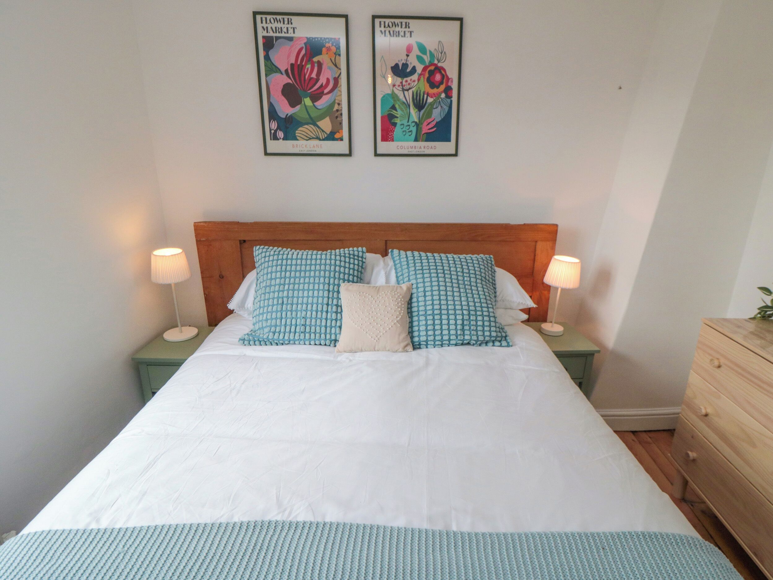 Een net opgemaakt bed met een witte dekbedovertrek en blauw gestreepte kussens, aan weerszijden voorzien van nachtkastjes met lampen, in een kamer met een houten hoofdbord en kunstwerken in lijsten aan de muur.