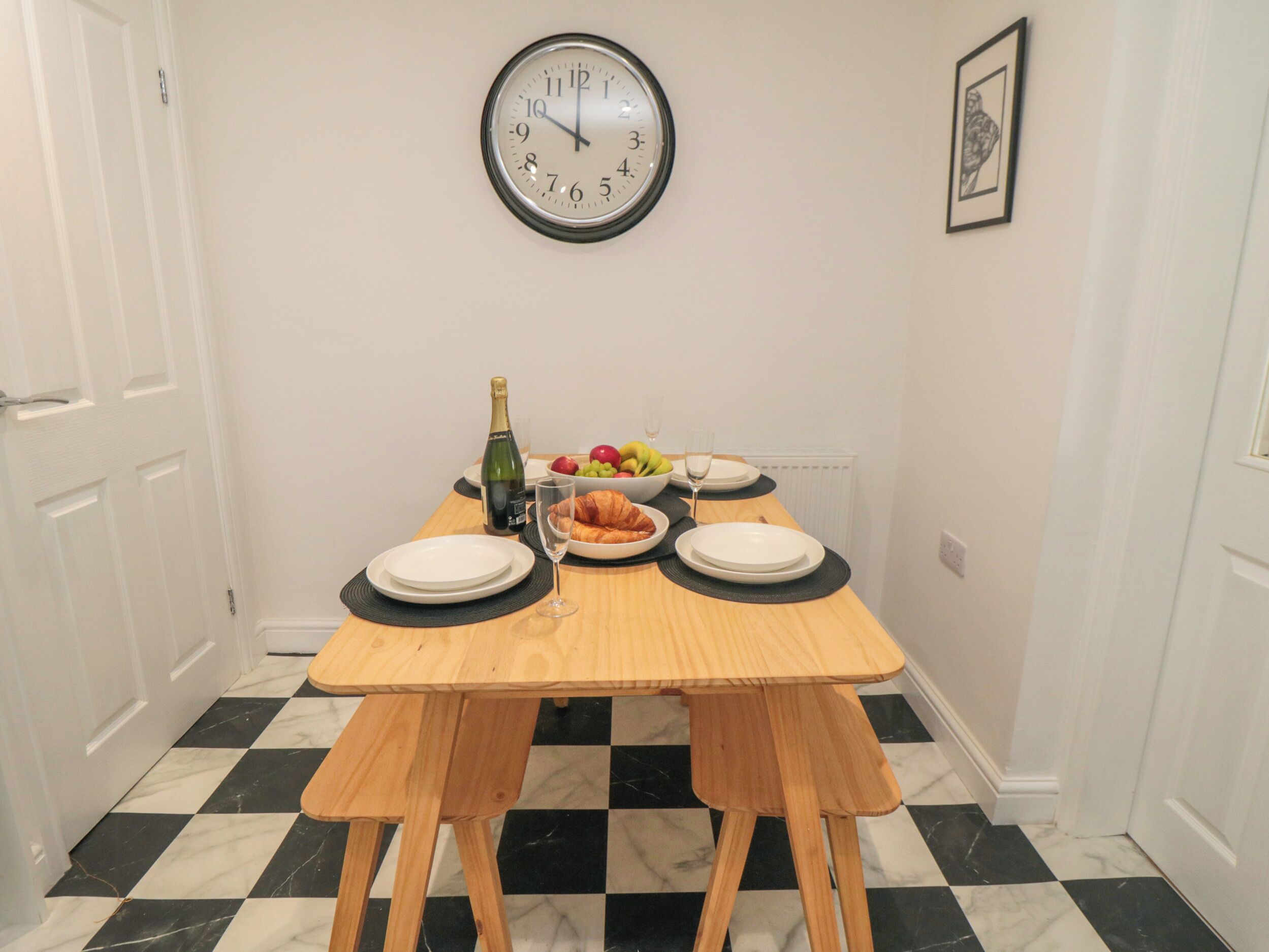 A dining area with a wooden table, black and white checkered floor, a wall clock, and a framed picture.