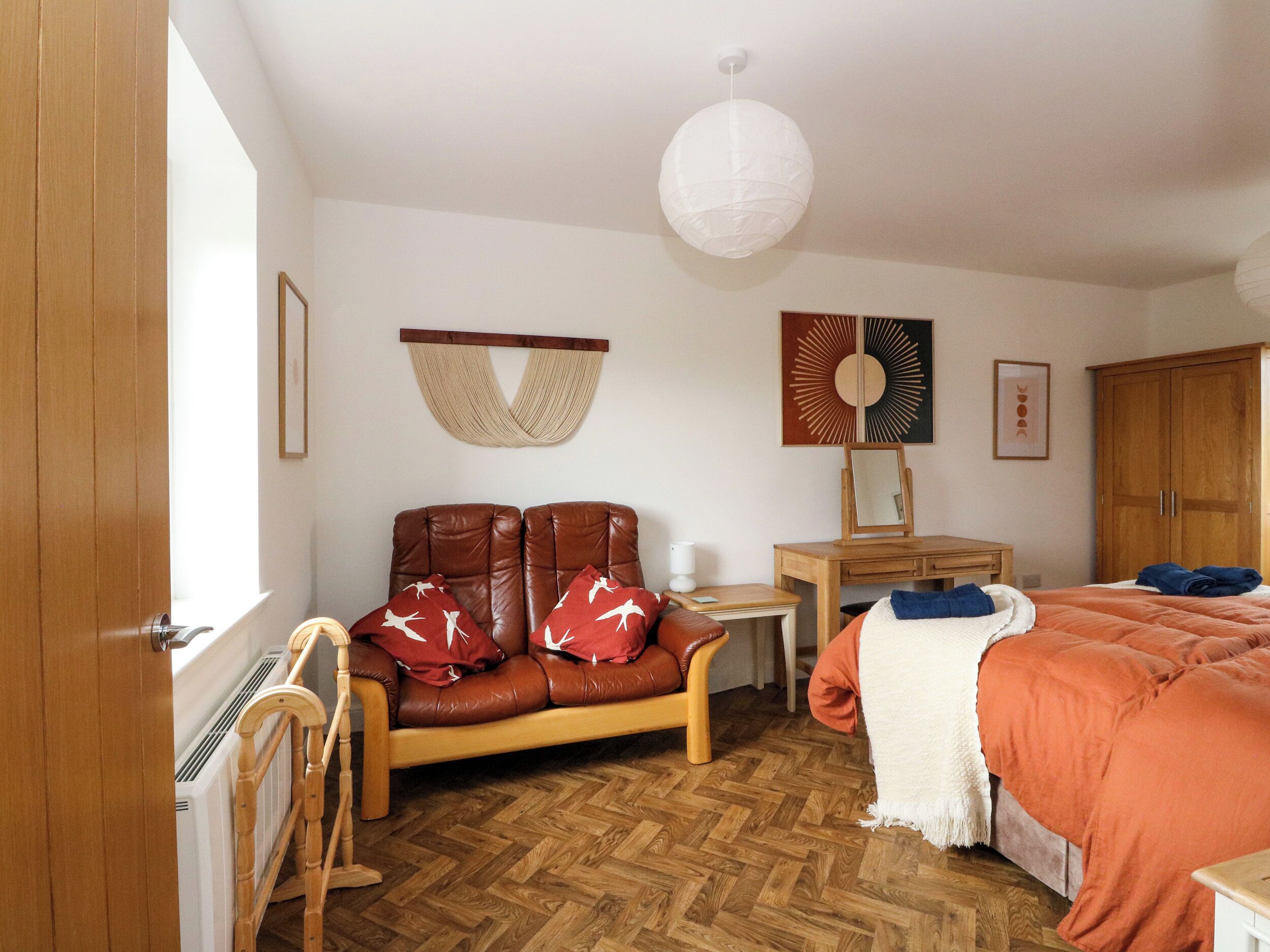 A bedroom with a wooden floor, a brown leather sofa, a wooden desk, a bed with an orange blanket, and a white wall hanging.