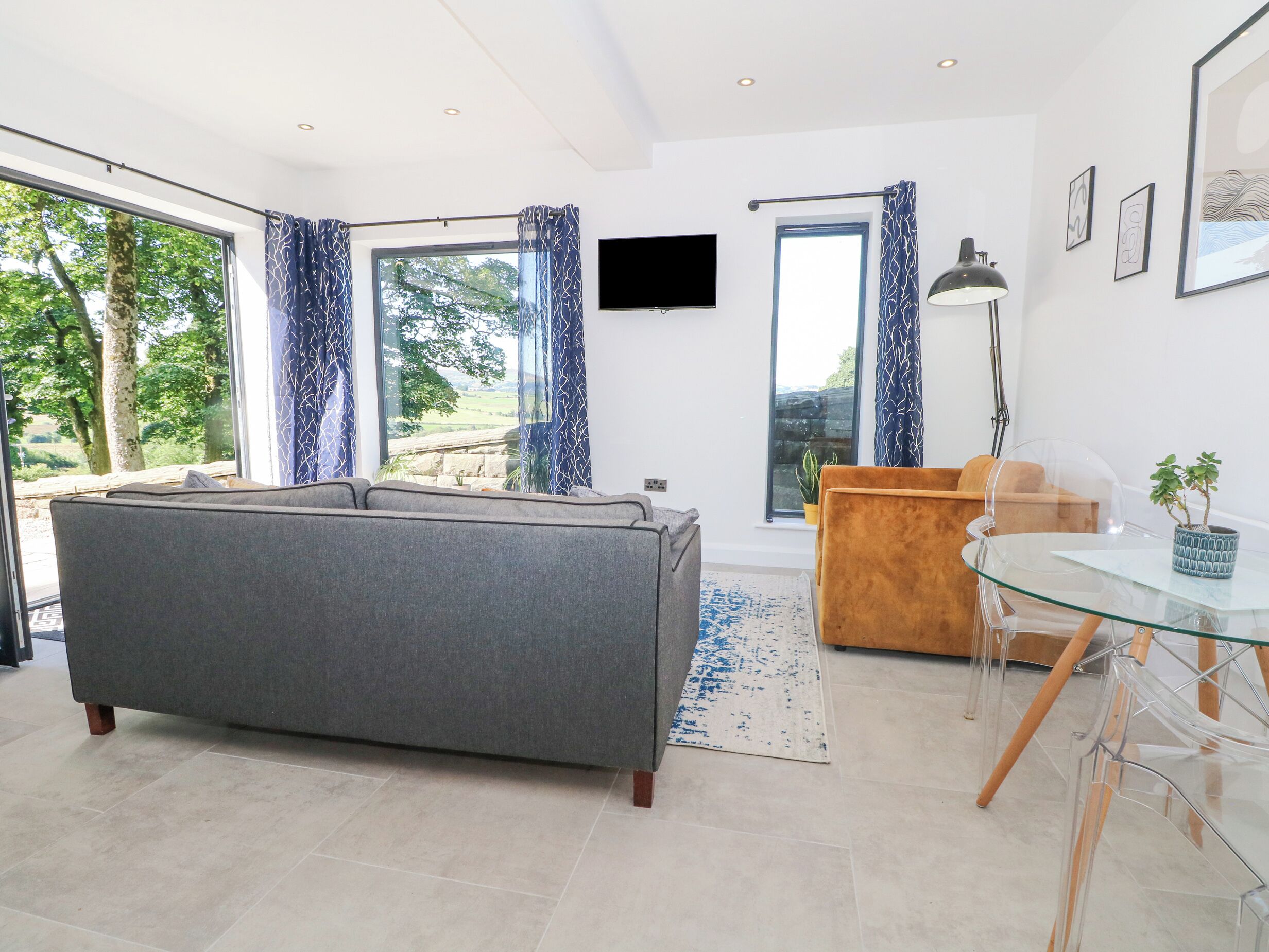 Una sala de estar moderna con un sofá gris, una mesa de comedor de vidrio con sillas, una mesita de madera y un televisor montado en la pared.