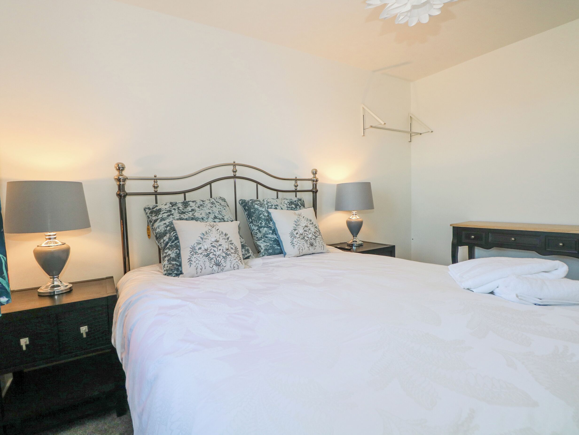 A neatly made bed with white bedding and two decorative pillows, flanked by two bedside tables with lamps, in a simple room with a plain wall and a ceiling light fixture.