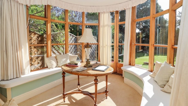 Een gezellige kamer met een ronde houten tafel, een bank met kussens en grote ramen met uitzicht op de tuin.