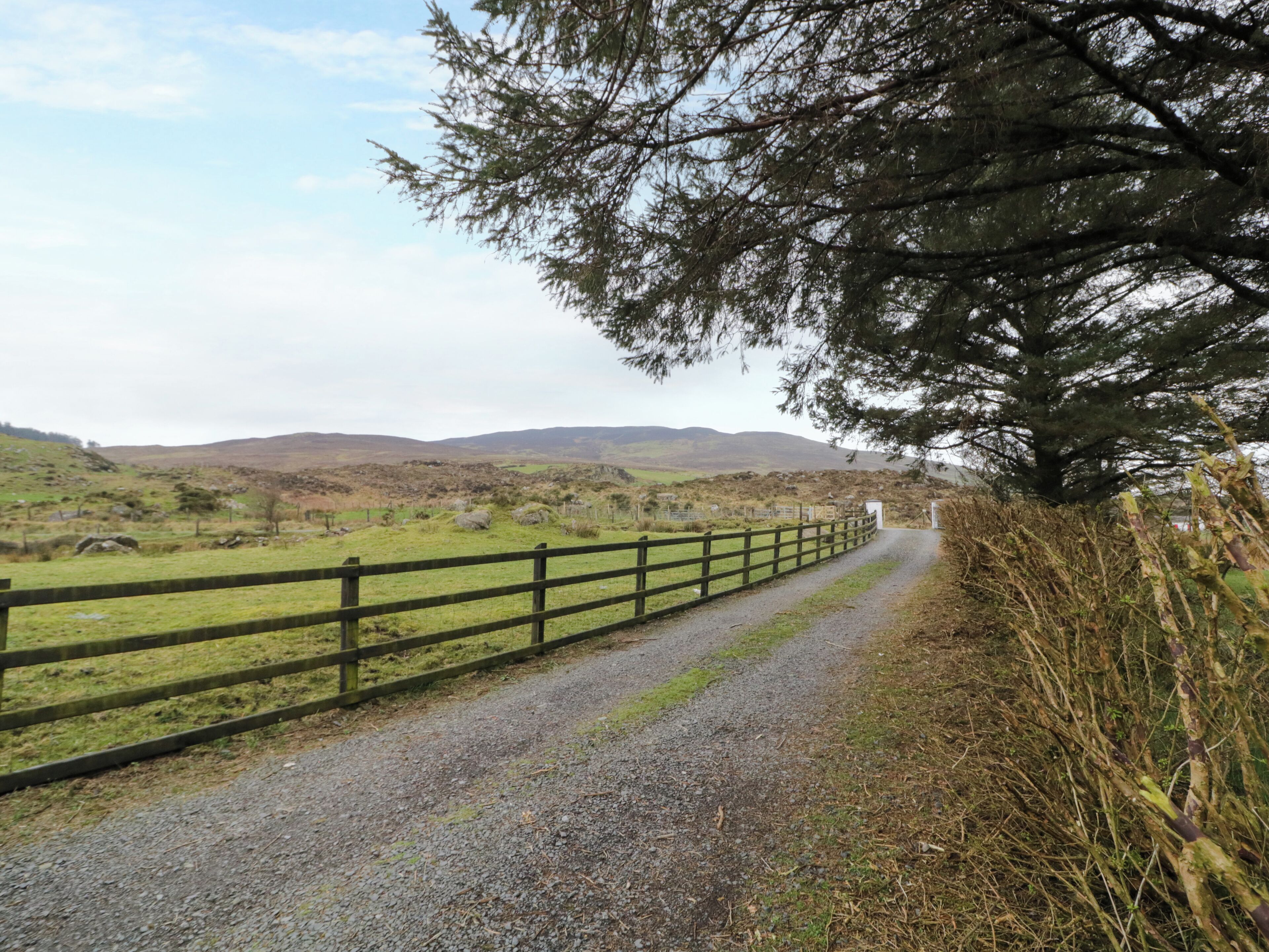A gravel path bordered by a wooden fence, leading through a rural landscape with rolling hills and sparse vegetation.