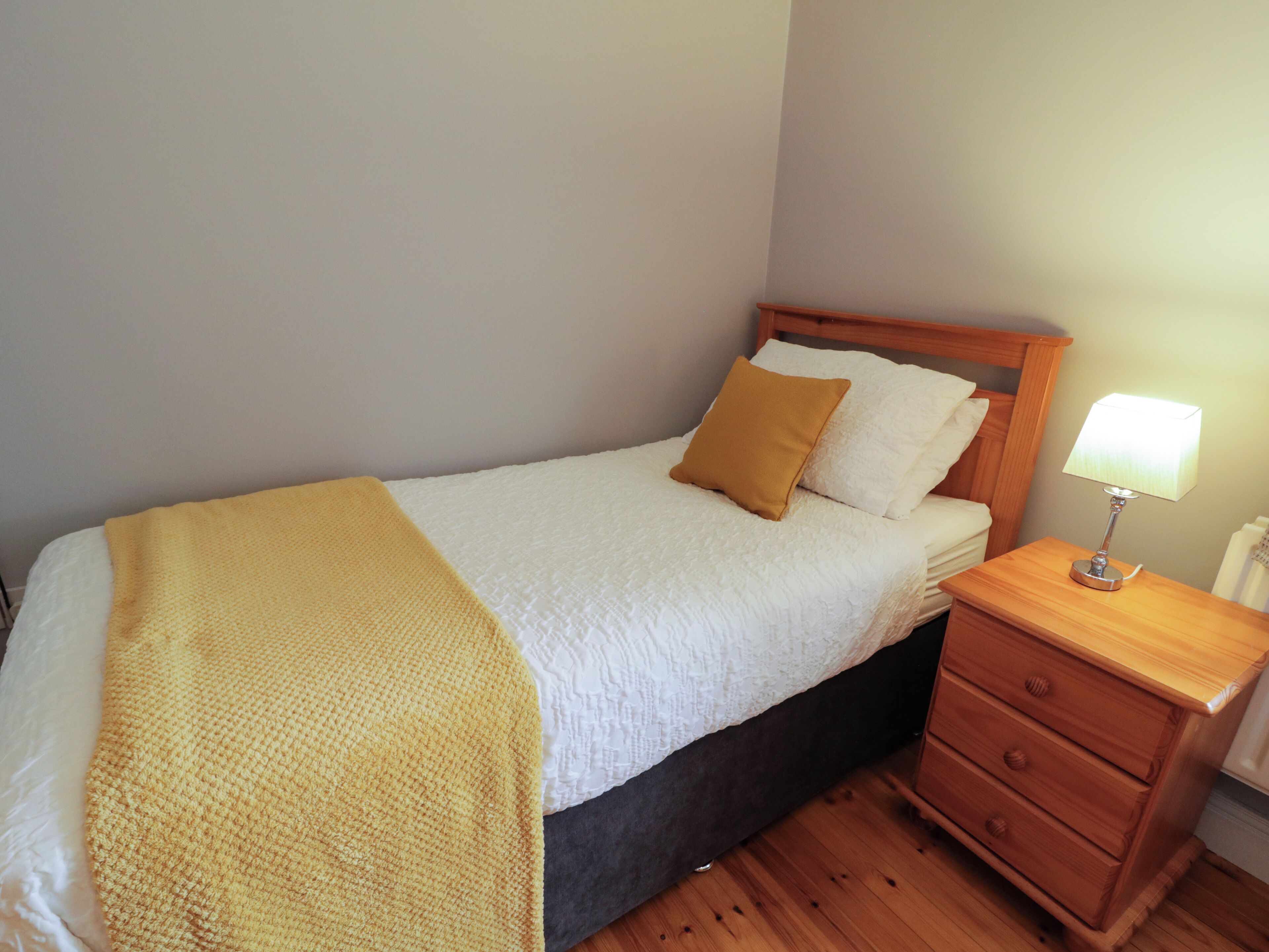 A neatly made bed with a white duvet and a yellow throw blanket, a wooden nightstand with a lamp, and a wooden dresser with three drawers.