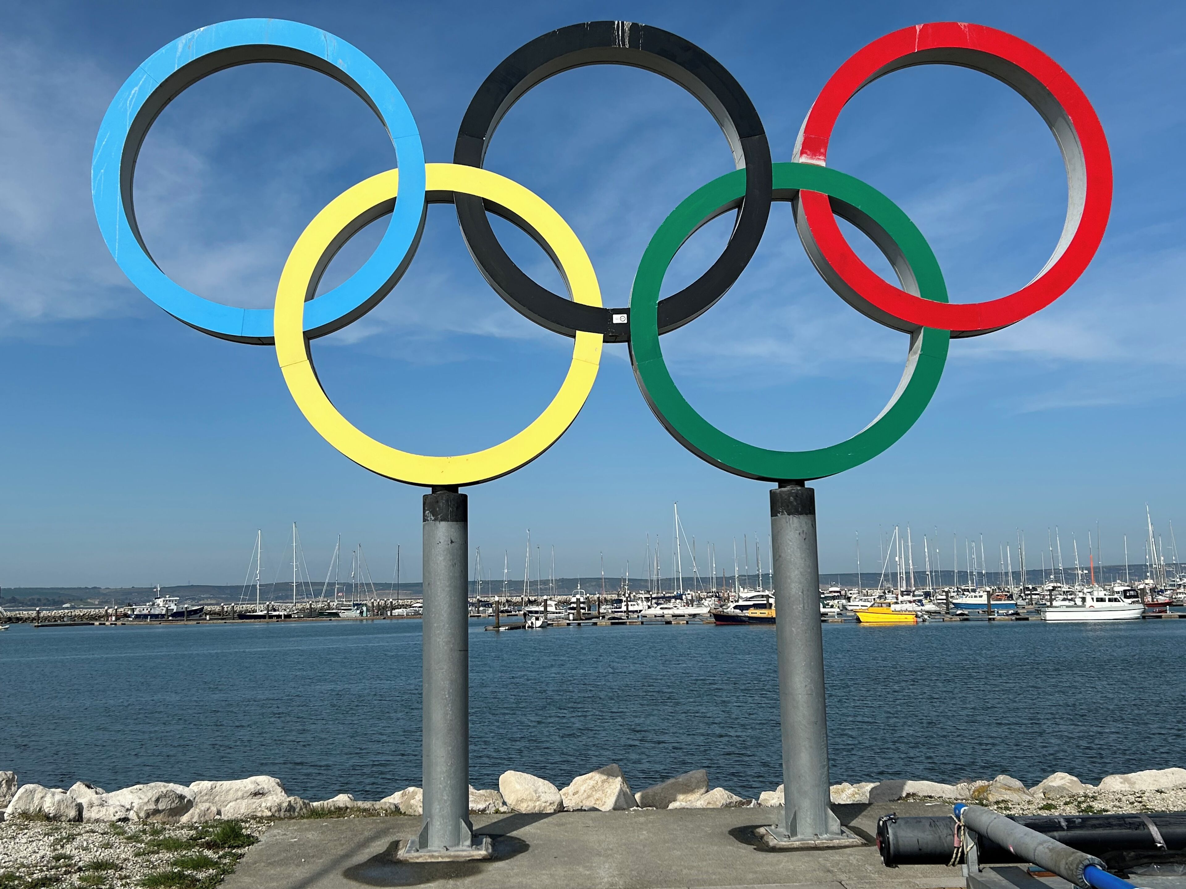 Olympic rings mounted on poles by a marina with boats in the background.