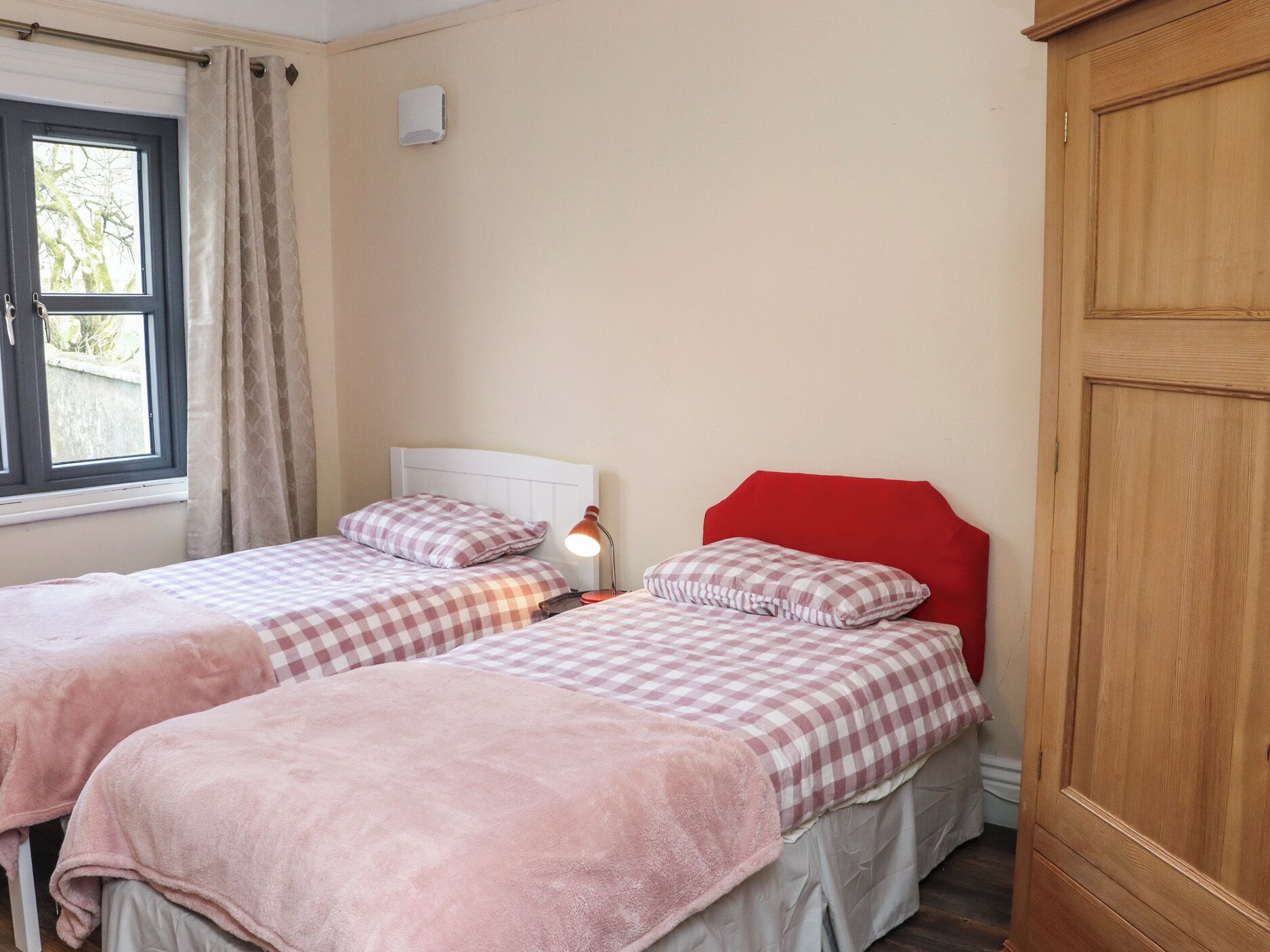 A room with two beds, each with a checkered bedspread and pink blankets, a bedside lamp, a wooden wardrobe, and a window with curtains.