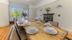 A dining room with a long wooden table set for a meal, flanked by chairs, with a fireplace and a view of greenery outside. - Fir Trees (Windermere)
