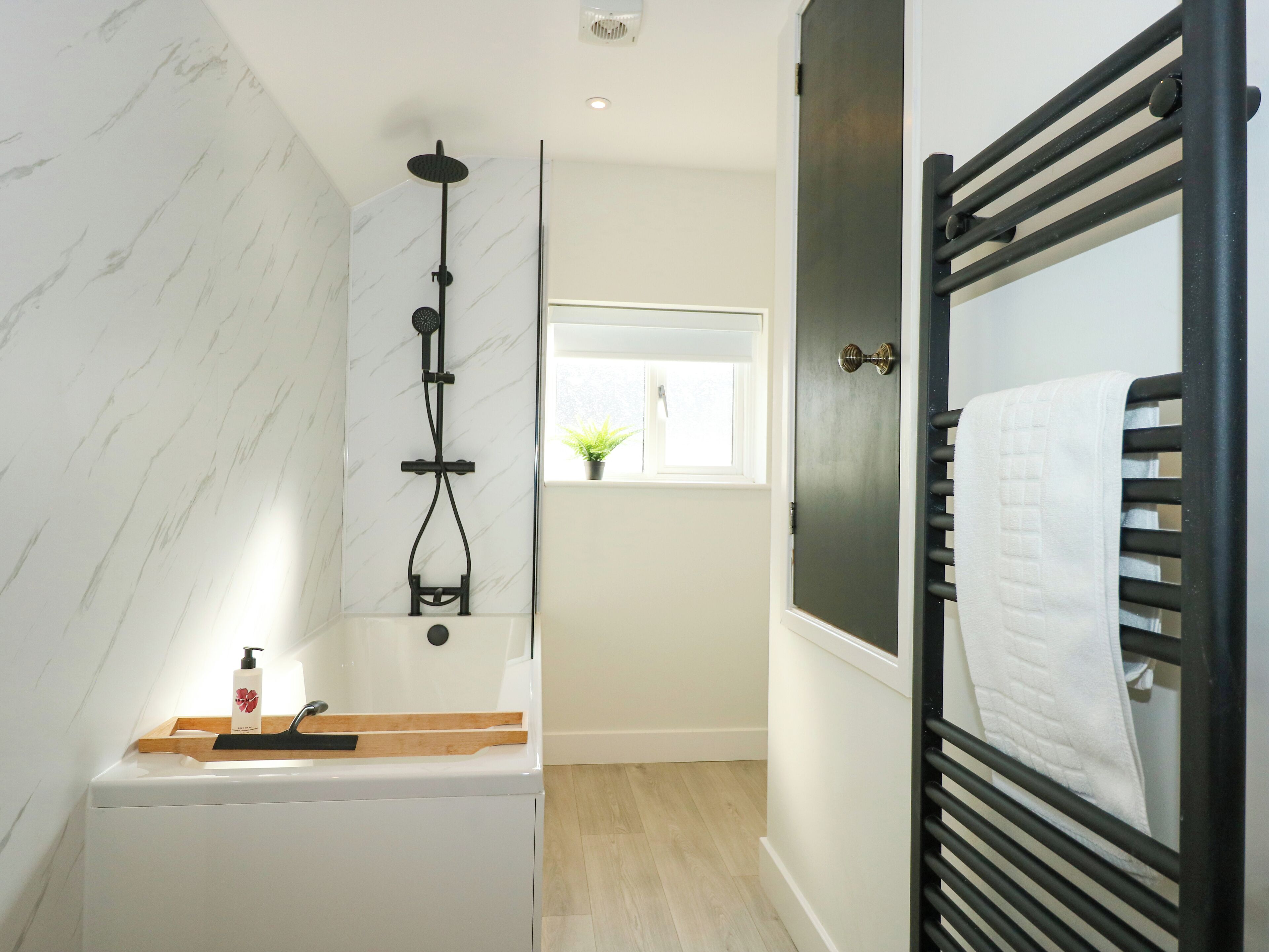 A modern bathroom with a marble wall, a black towel rack, a wooden tray with toiletries, and a window with a plant.