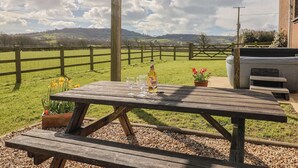 A wooden picnic table with a bottle and glasses, set against a rural backdrop with a fence and greenery. - Daisy Cottage (Bridport)