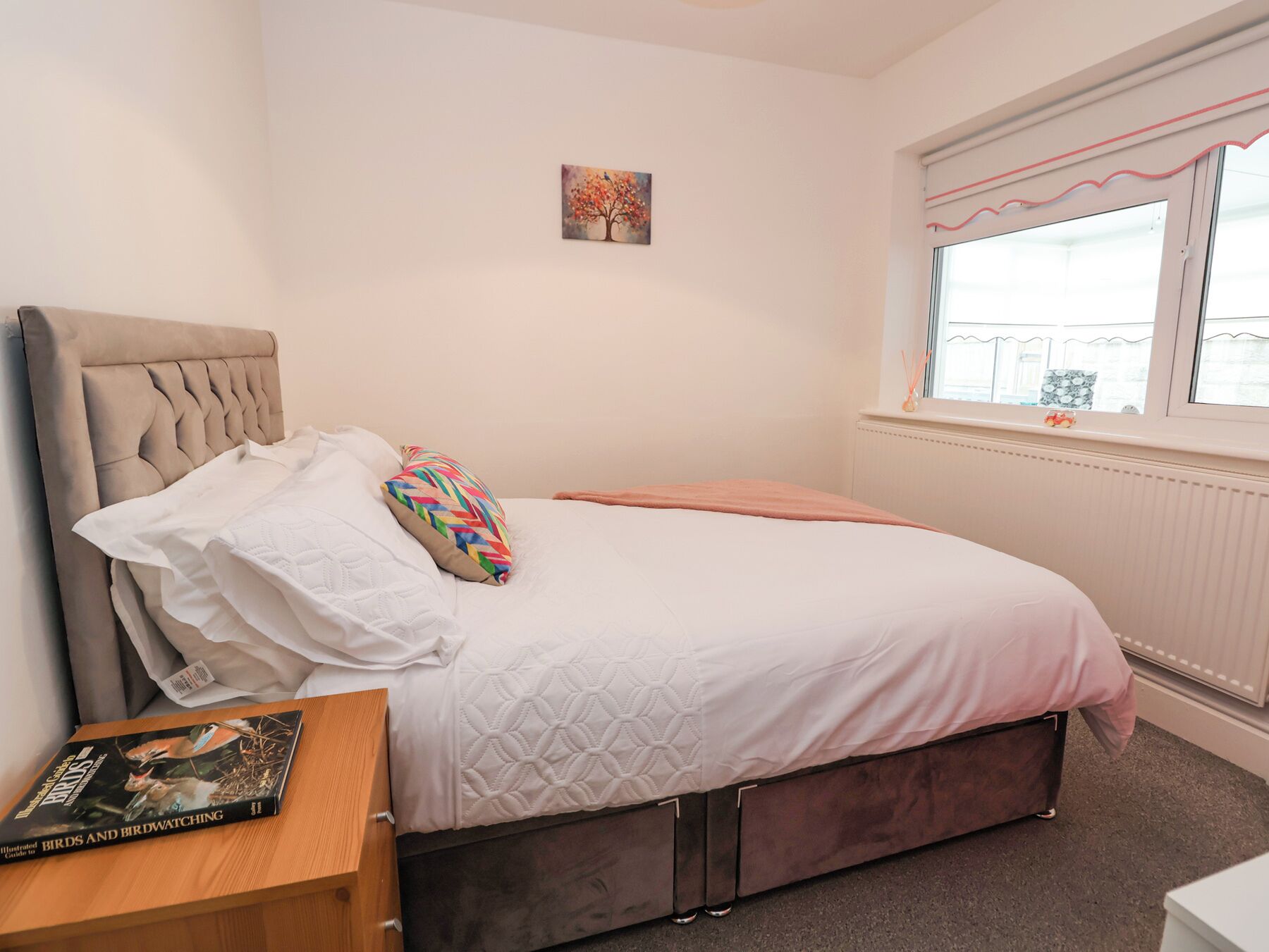 A bedroom with a bed, a window, a radiator, and a small table with a book.