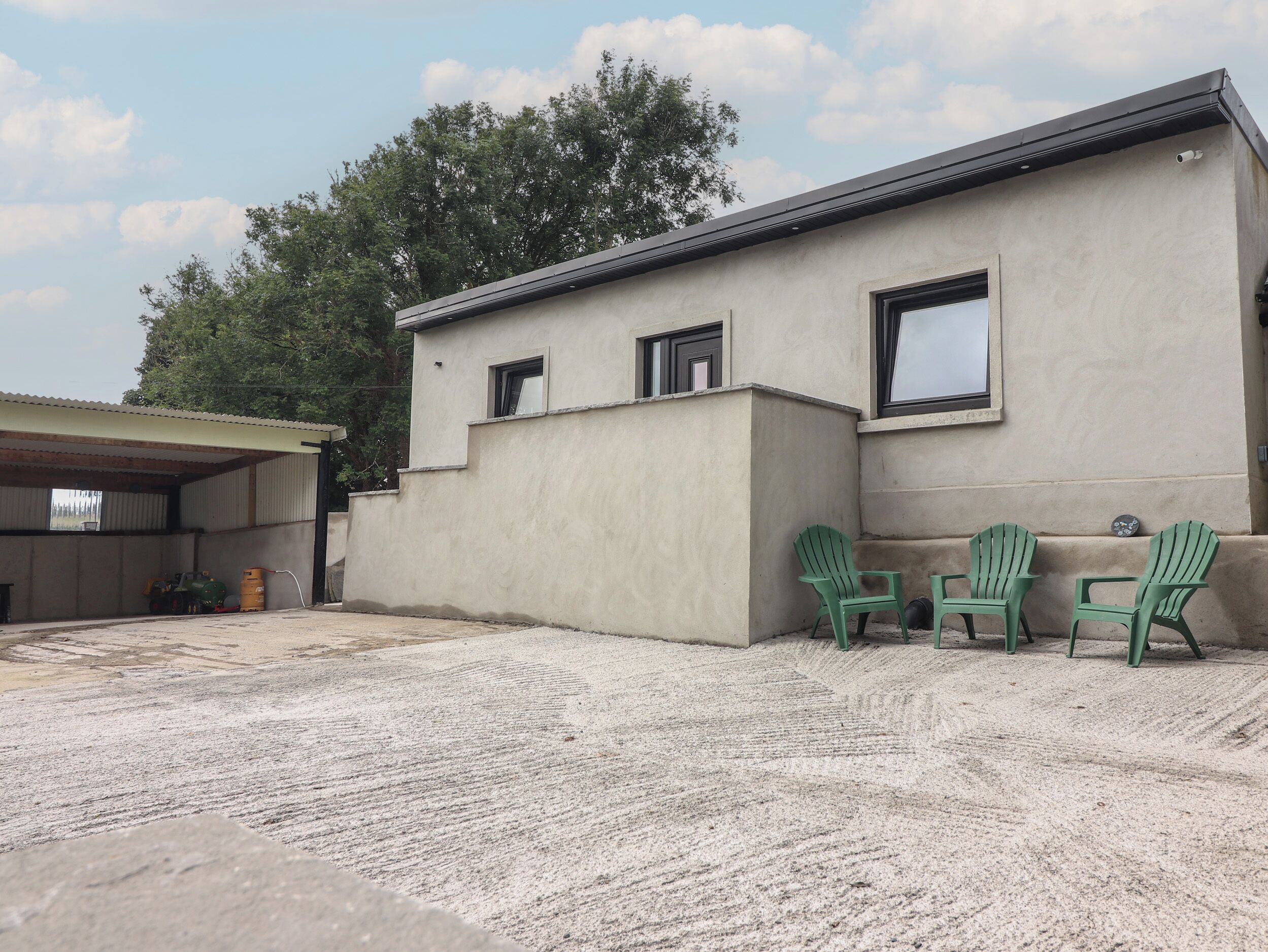 A building with a flat roof, a concrete facade, and a covered area with a roof. There are three green chairs outside.