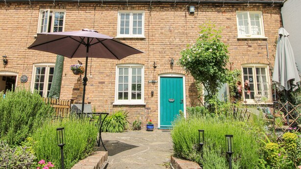 Une maison en briques avec une porte verte, une terrasse avec un parasol et un jardin où poussent différentes plantes.