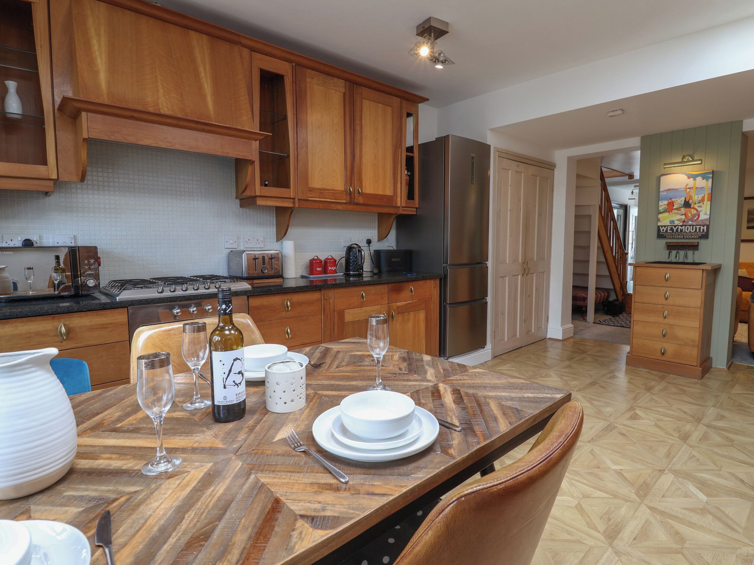 A kitchen with wooden cabinets, a stainless steel refrigerator, a dining table set with plates, glasses, and cutlery.