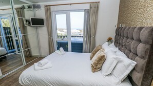 A neatly made bed with white linens and a grey headboard, a bedside table with a laptop, and a view of the outdoors through a sliding door. - Bay View (Redruth)