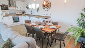 A modern kitchen with white cabinets, a wooden dining table set for four, and a grey sofa in the living area. - Bay View (Redruth)