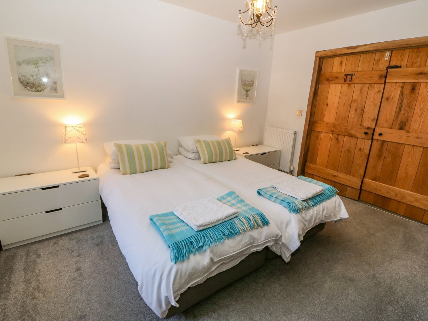 A double bed with white bedding and two pillows, a bedside table with a lamp, a wooden wardrobe, and two wall-mounted lamps.