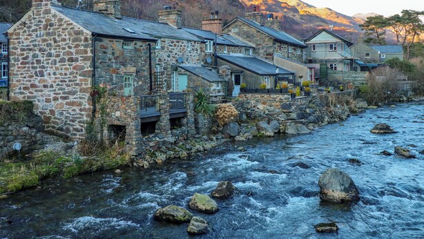 Un edificio in pietra con un fiume di fronte, circondato dalle montagne.