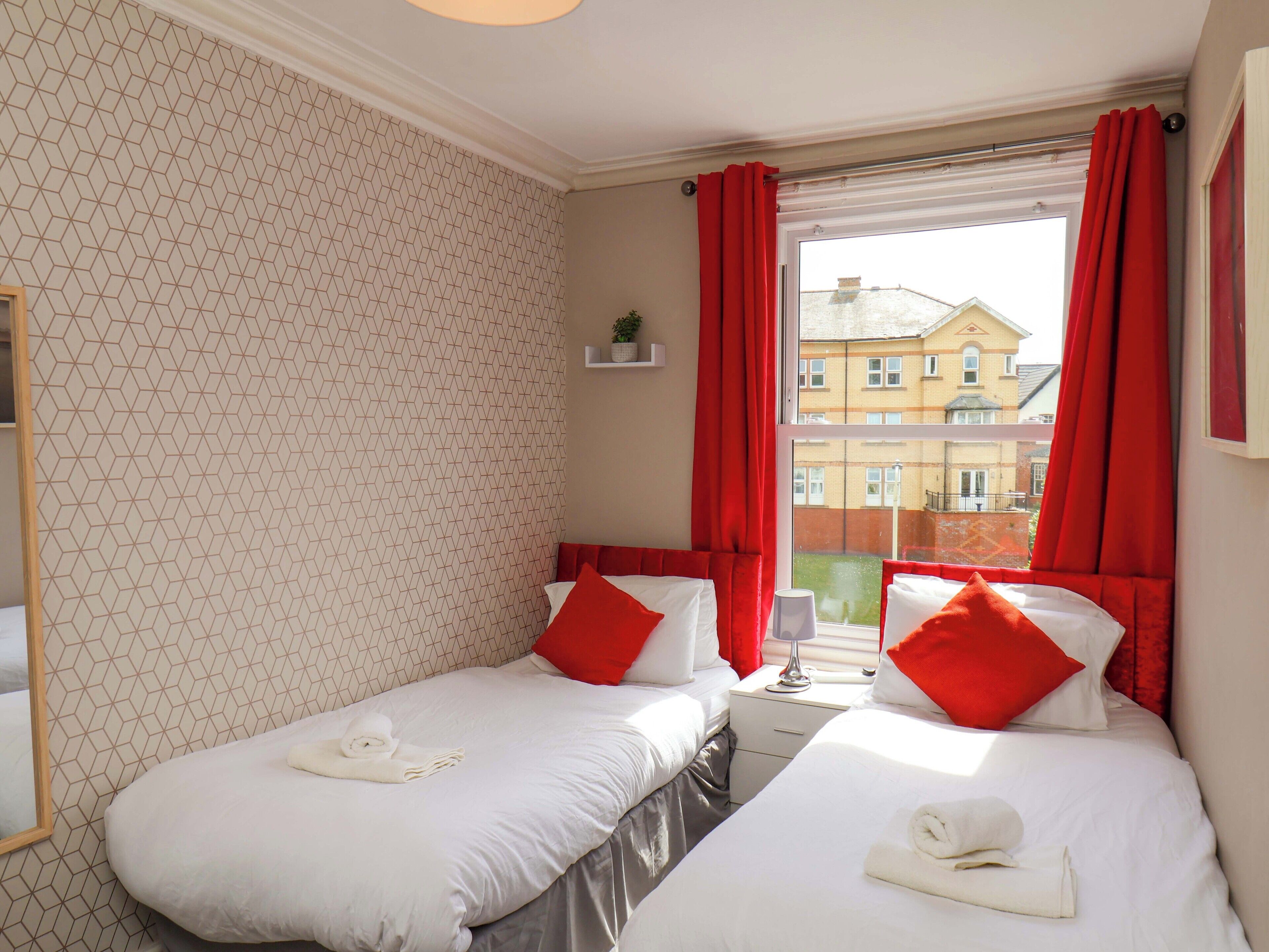 Ett hotellrum med två separata sängar, röda gardiner, ett fönster med utsikt över byggnader och en vägg med ett geometriskt mönster.