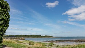 Un sentier pavé mène à une plage, sous un ciel dégagé, avec des collines qui se dessinent à l’horizon.