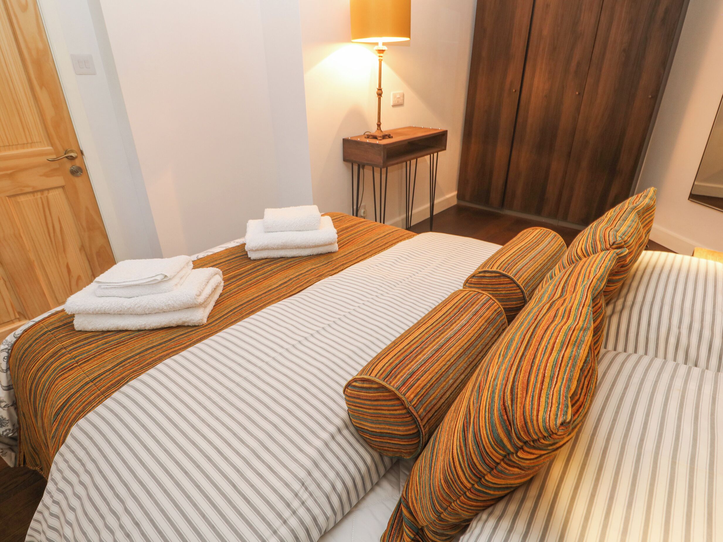 Un lit bien fait, avec une literie à rayures et des coussins à rayures enroulés, une table de chevet avec une lampe et une porte en bois, le tout dans une chambre d'hôtel.