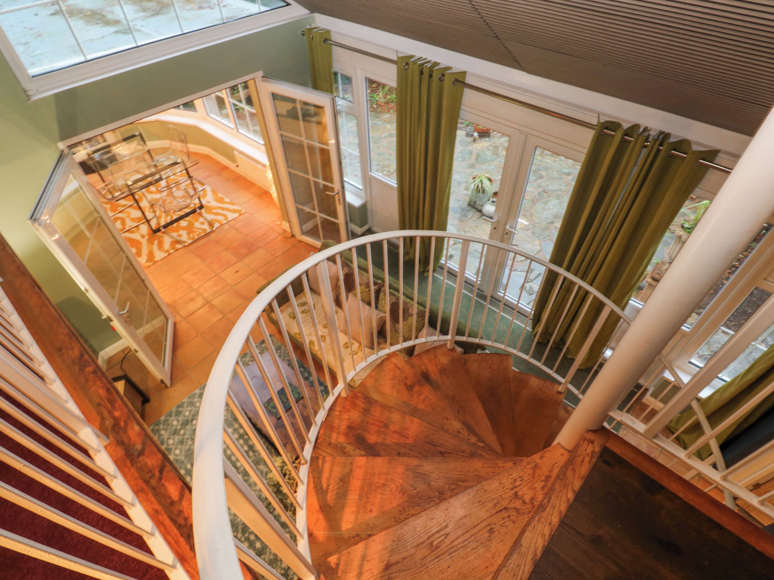 A spiral staircase with a wooden finish, leading to a room with a view of a garden and a dining area with a table and chairs.