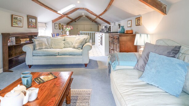 A spacious loft with a high ceiling, exposed wooden beams, a light blue sofa, a white sofa, a wooden coffee table, and a kitchen area in the background.