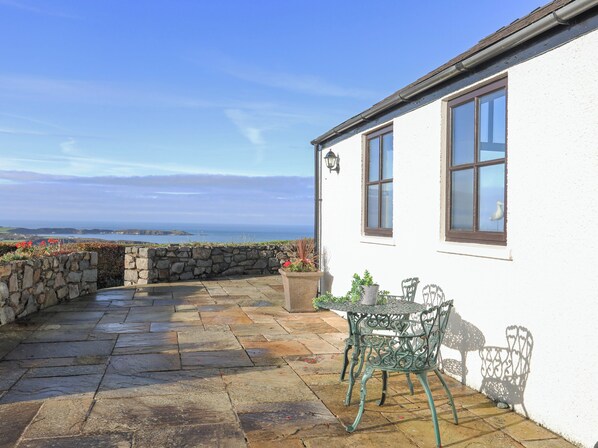 A stone patio with a table and chairs, a white building with windows, and a view of the sea. - Tai Ffolt (Pwllheli)