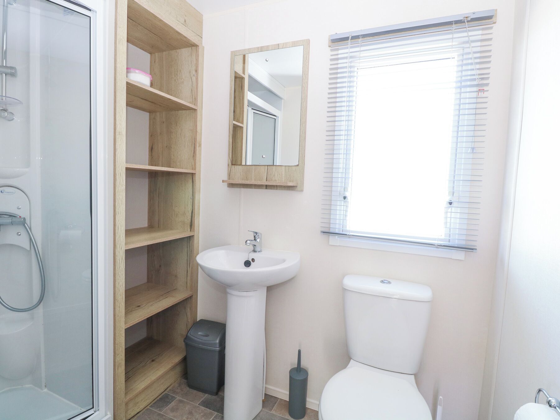 A bathroom with a glass shower enclosure, a white sink, a toilet, and a window with blinds.