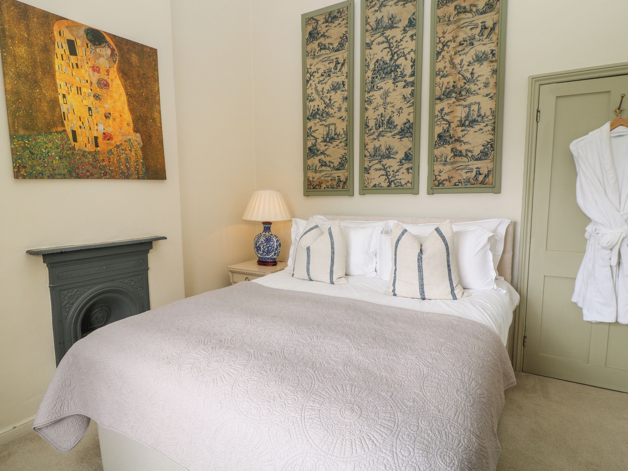 A bedroom with a large bed, a nightstand with a lamp, a fireplace, and artwork on the walls.