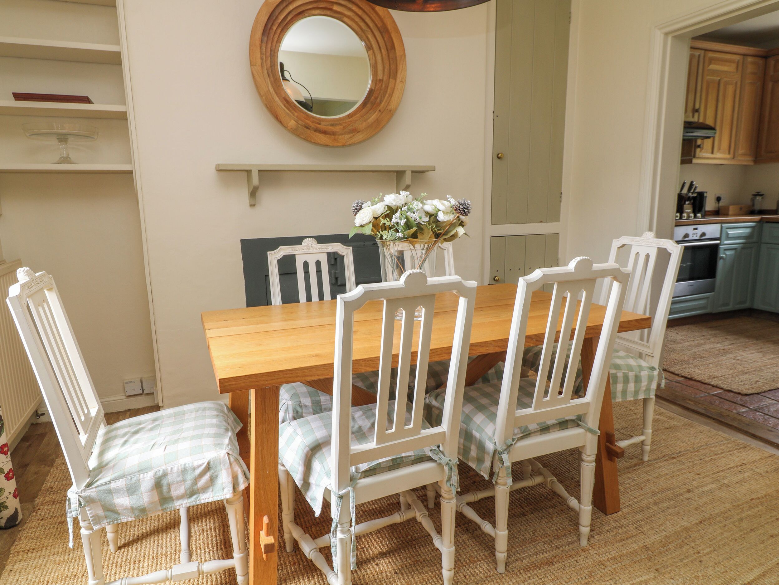 A dining area with a wooden table and white chairs, a mirror on the wall, and a kitchen in the background.