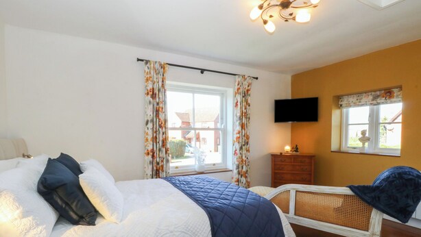 A bedroom with a bed, a dresser, a television, and a window with curtains. - Cobble Cottage (Fakenham)