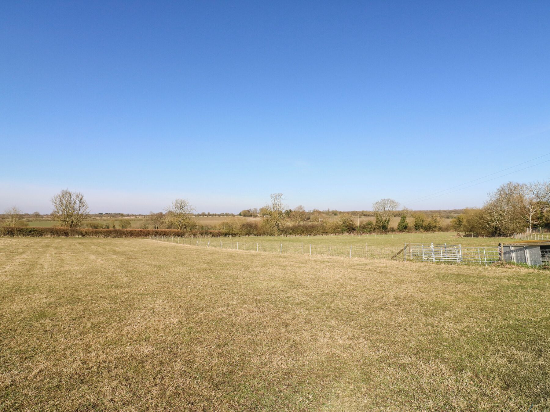 A wide, open grassy field with a metal fence in the foreground and trees in the background under a clear blue sky.