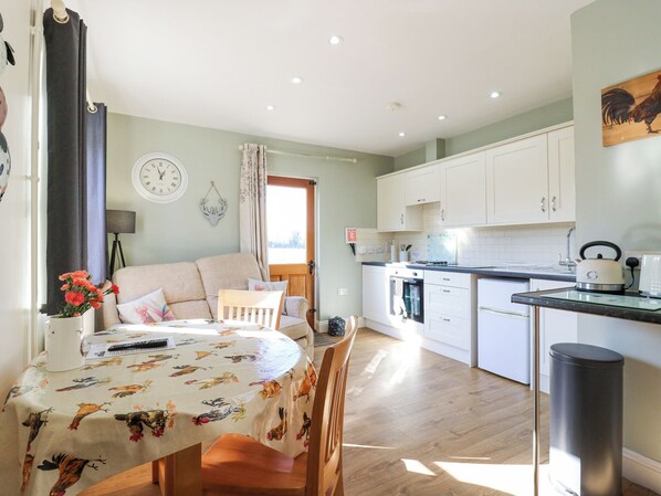 A cozy living room with a sofa, a dining table with a floral tablecloth, and a kitchen with white cabinets and a kettle on the counter. - B&b 1 (Saffron Walden)