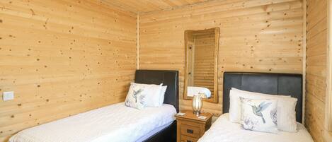 A wooden cabin room with two beds, each with a pillow featuring a bird design. There is a nightstand with a lamp between the beds.