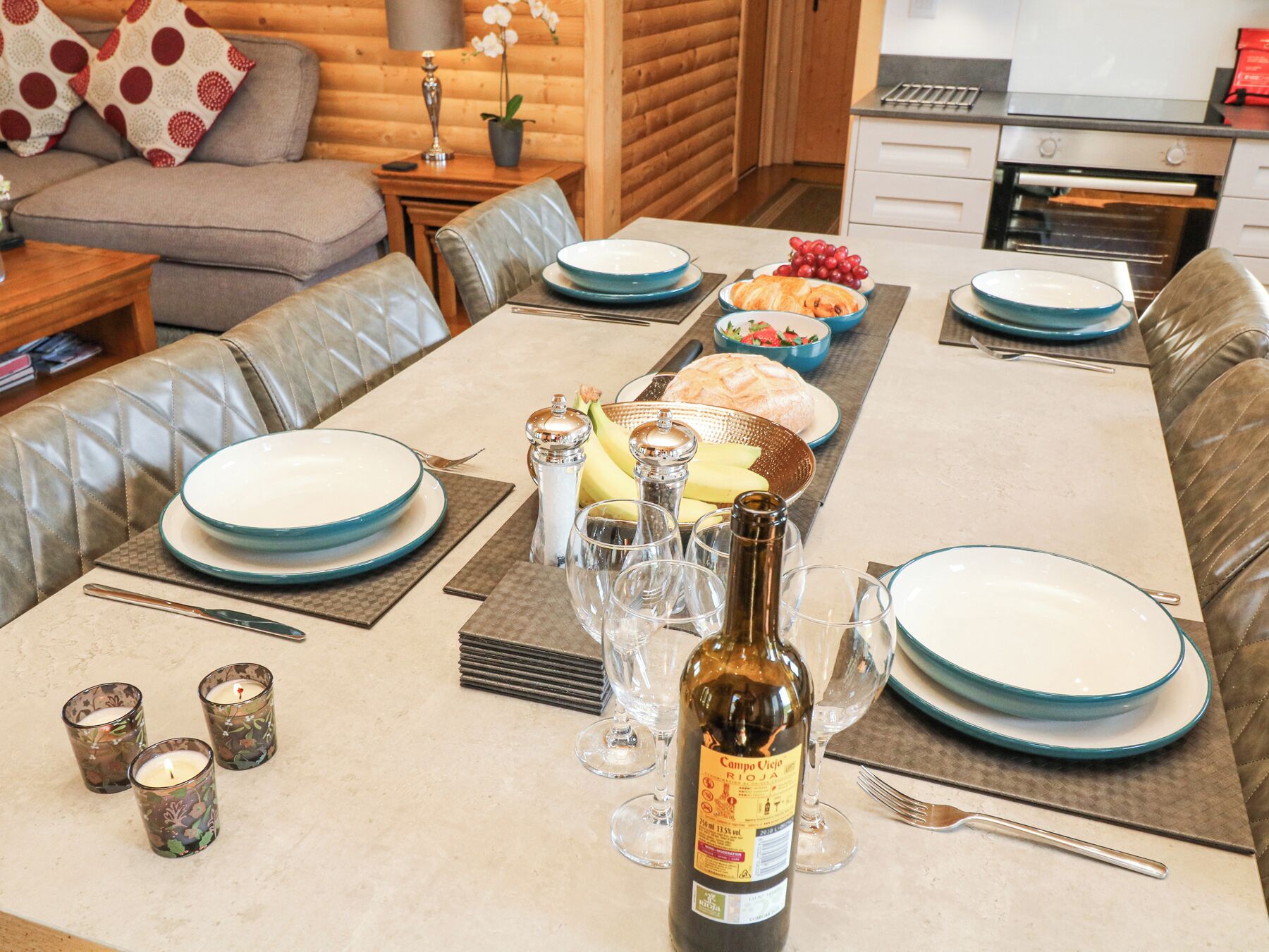 A dining table set with plates, glasses, and a bottle of wine, surrounded by leather sofas and a kitchen area in the background.