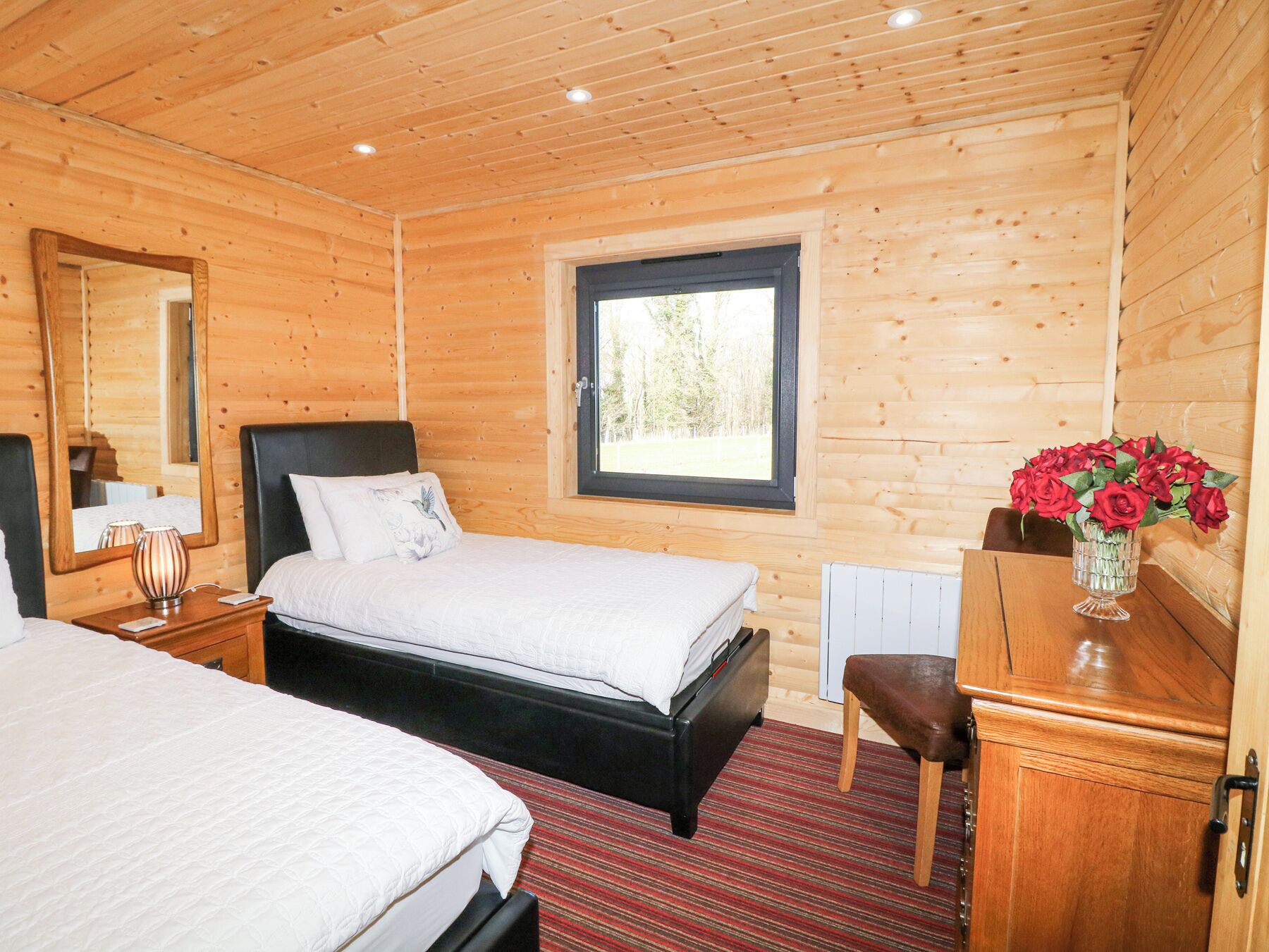 A wooden cabin bedroom with a bed, a nightstand, a dresser, a mirror, and a window with a view of trees.