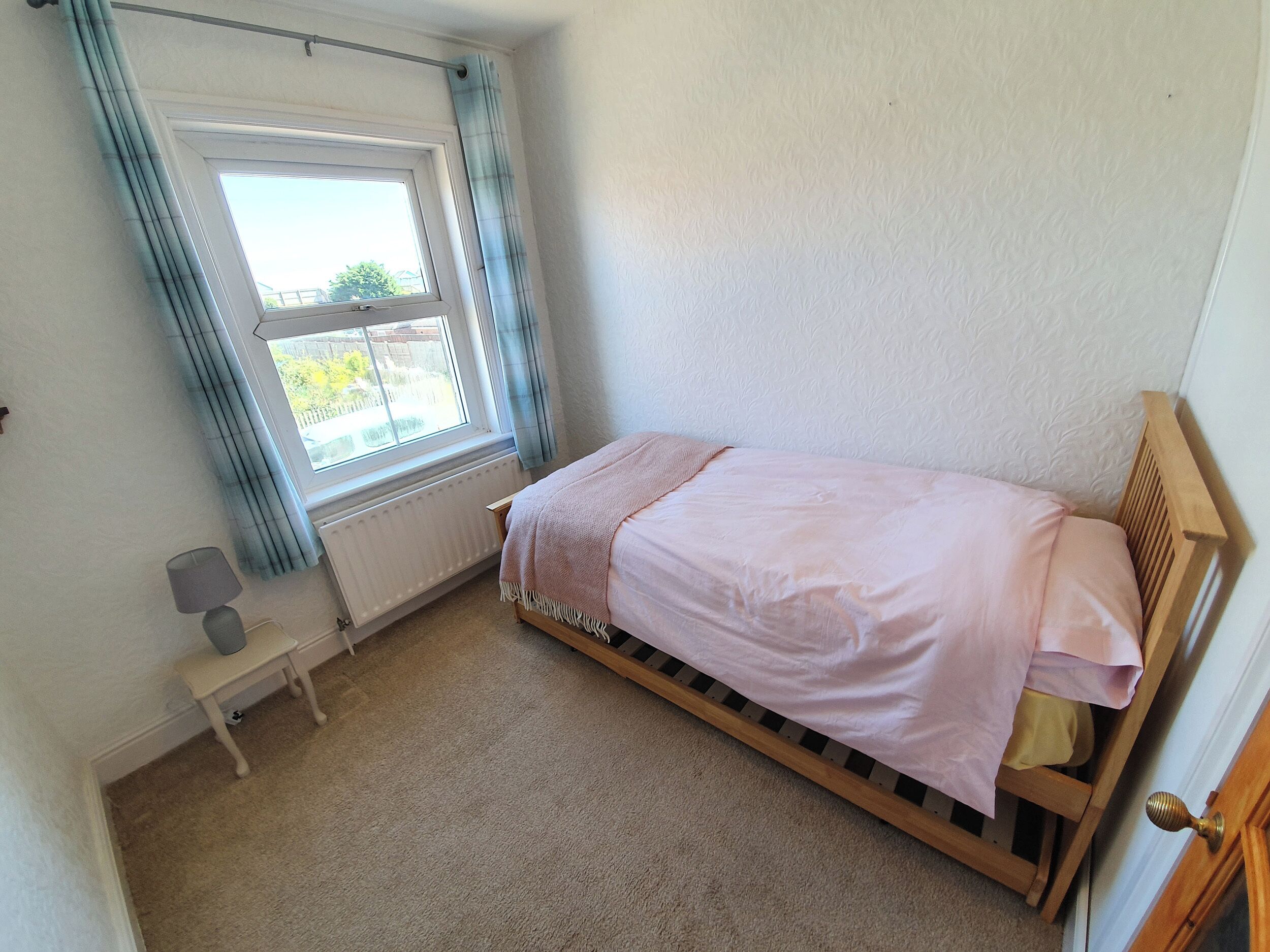 A small bedroom with a single bed, a radiator, a window with curtains, a bedside table, and a lamp.