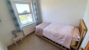 A small bedroom with a single bed, a radiator, a window with curtains, a bedside table, and a lamp.