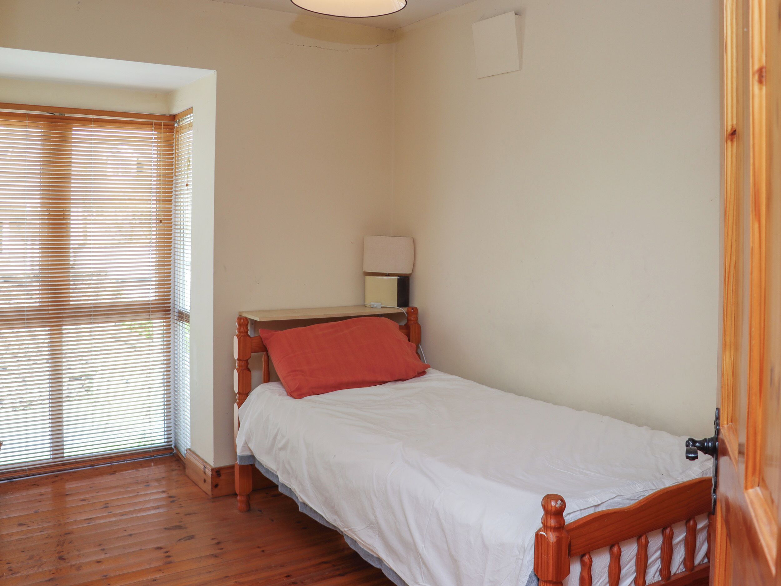A small, single-bed room with wooden flooring, a wooden headboard, and a red pillow.
