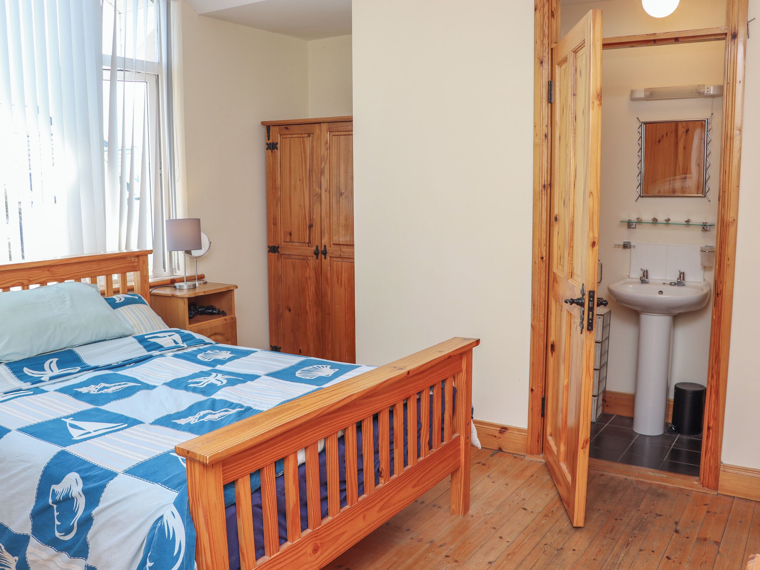 A bedroom with a wooden bed, a nightstand with a lamp, a wardrobe, and a bathroom with a sink and mirror.