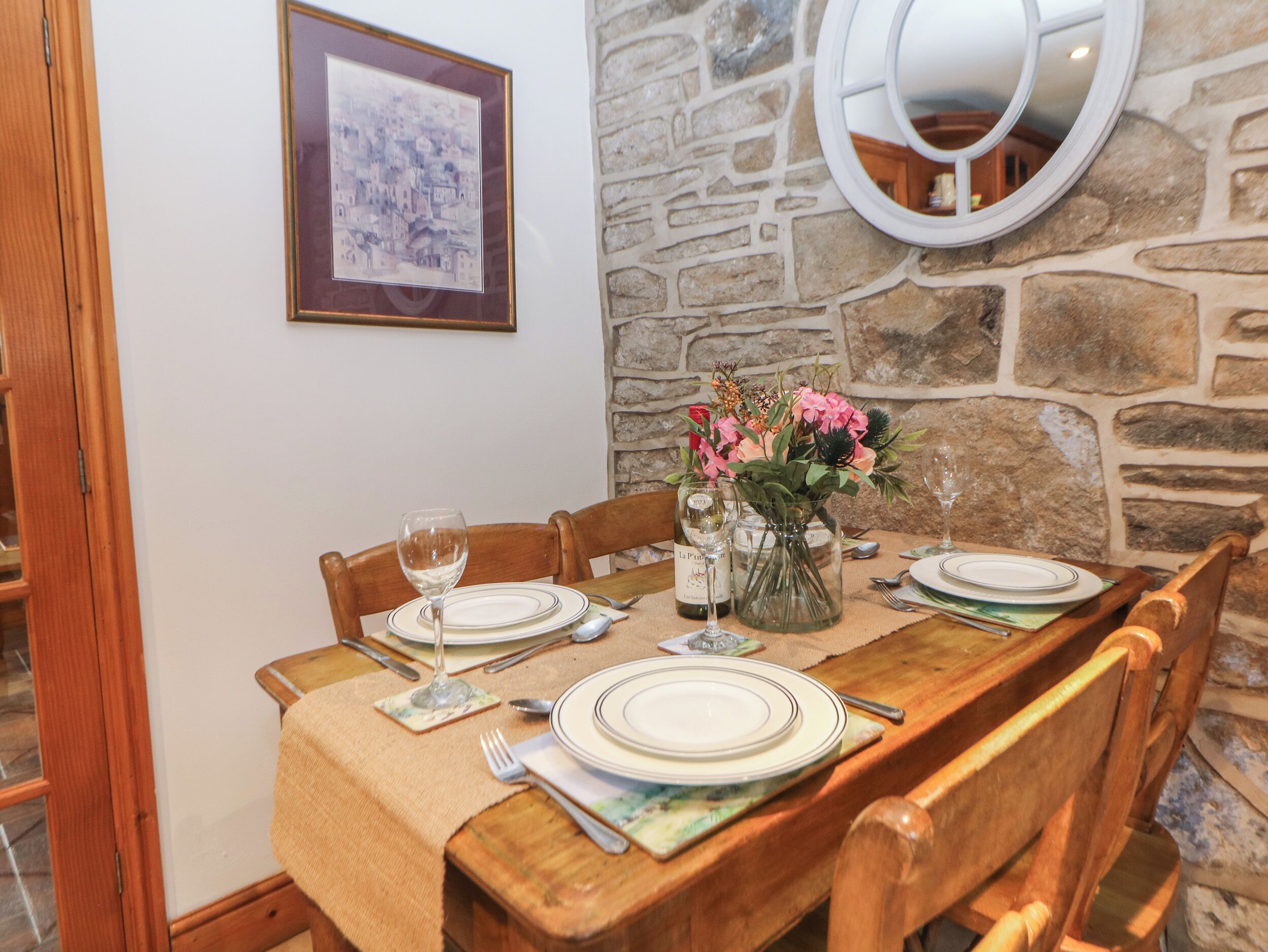 A dining table set with plates, cutlery, and a wine bottle, surrounded by wooden chairs, with a stone wall and a framed picture in the background.