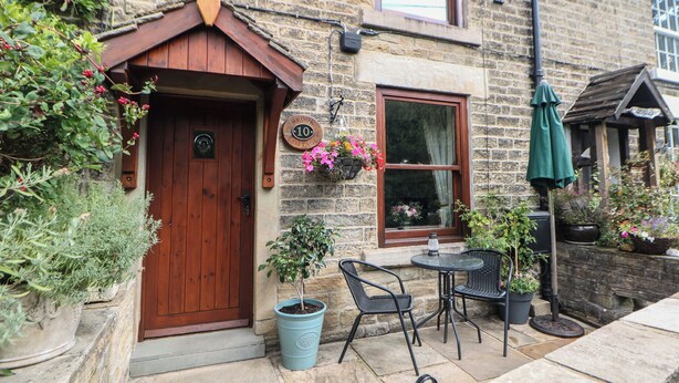 A stone cottage with a wooden door, a window, and a sign indicating it is number 10. There are potted plants and outdoor furniture. - Brook Cottage (High Peak)