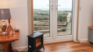A room with a wooden floor, a small black stove, a table with a lamp and glasses, and a view of a garden through a glass door. - Meadow Cottage (Scarborough)