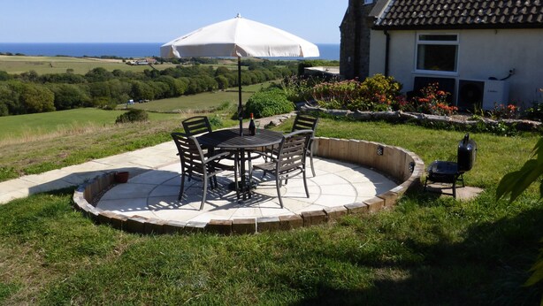 A patio with a table and chairs, a white umbrella, and a view of the sea. - Meadow Cottage (Scarborough)