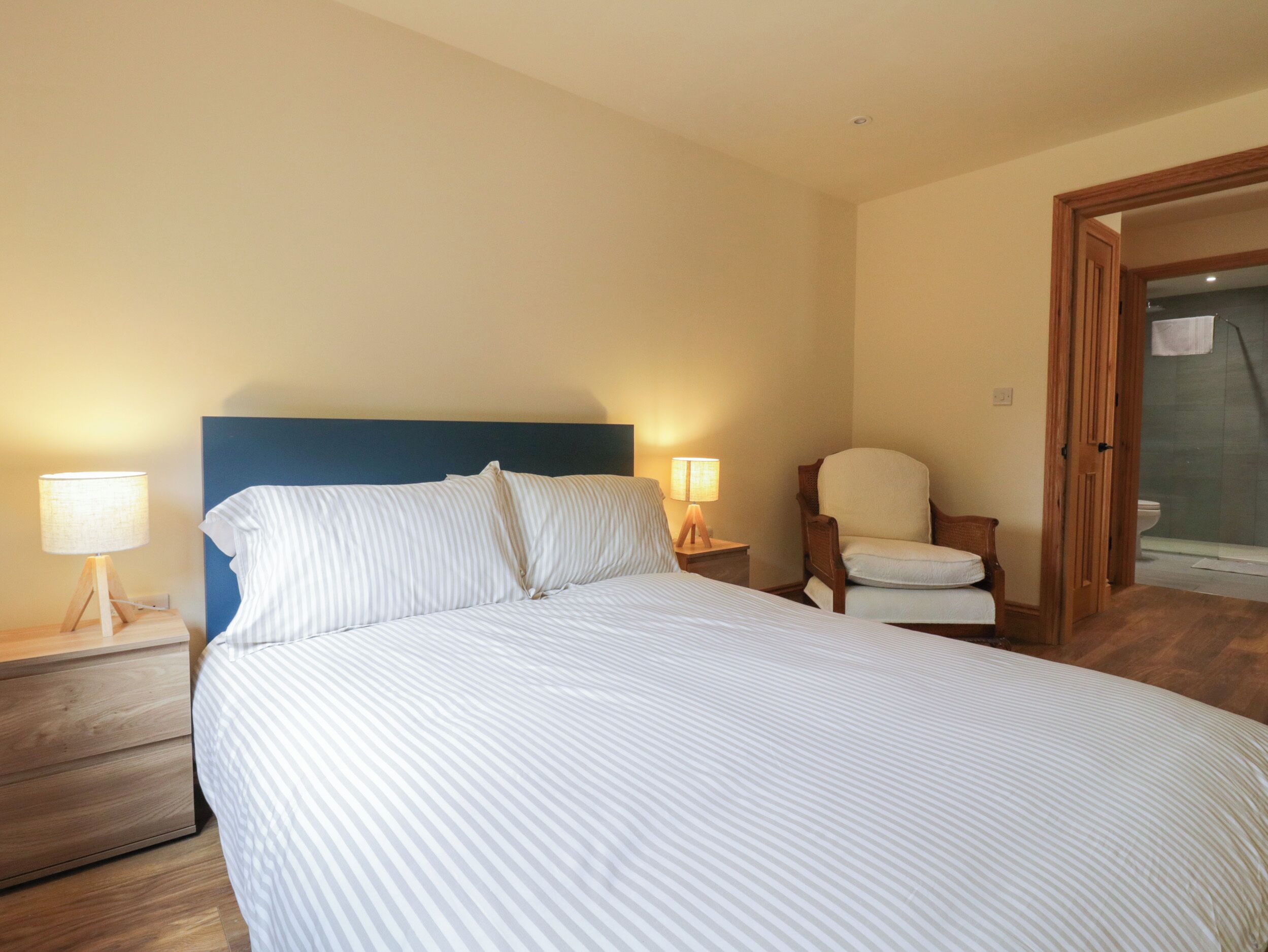 Una cama bien hecha con ropa de cama a rayas, dos lámparas de noche, una mesita de noche de madera, una silla de madera y una puerta de madera que da a un baño.