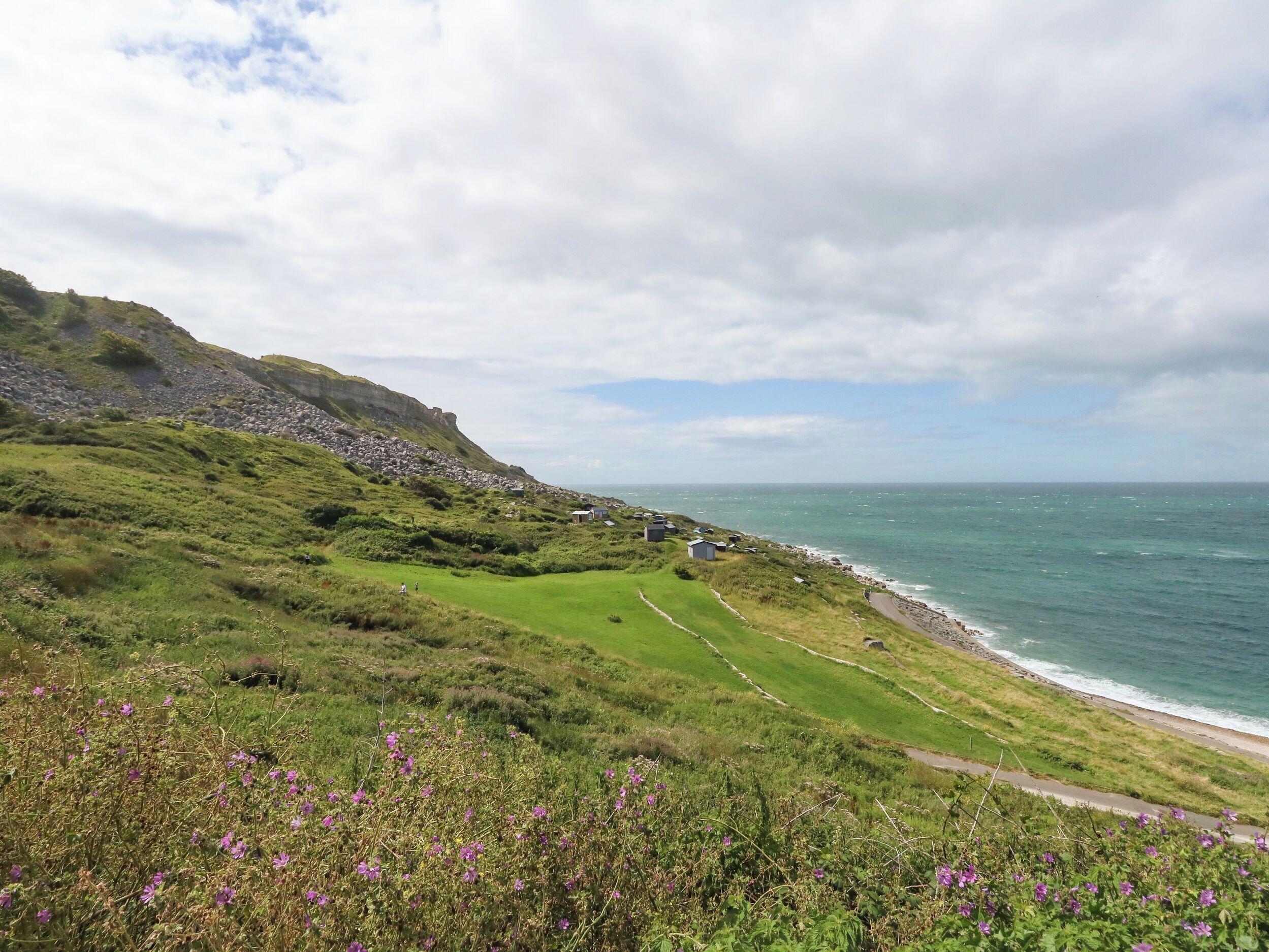 A coastal landscape with a grassy path leading to a beach, overlooking the sea and a rocky cliff.