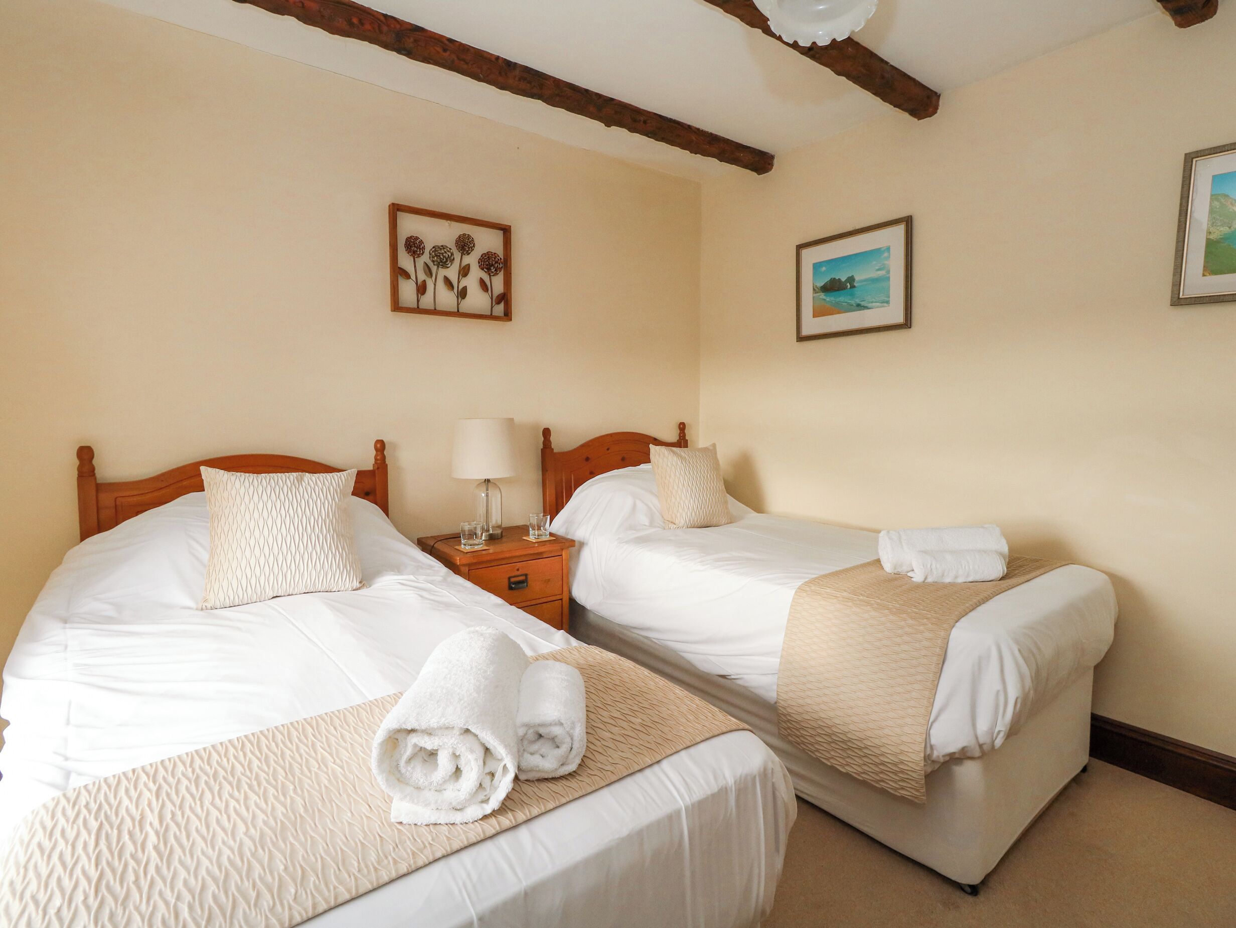 Een hotelkamer met twee eenpersoonsbedden, elk met een houten hoofdbord, witte beddengoed en opgerolde handdoeken. Bij elk bed staat een nachtkastje met een lamp en glazen. De kamer heeft een houten plafond en een lijst met een foto aan de muur.