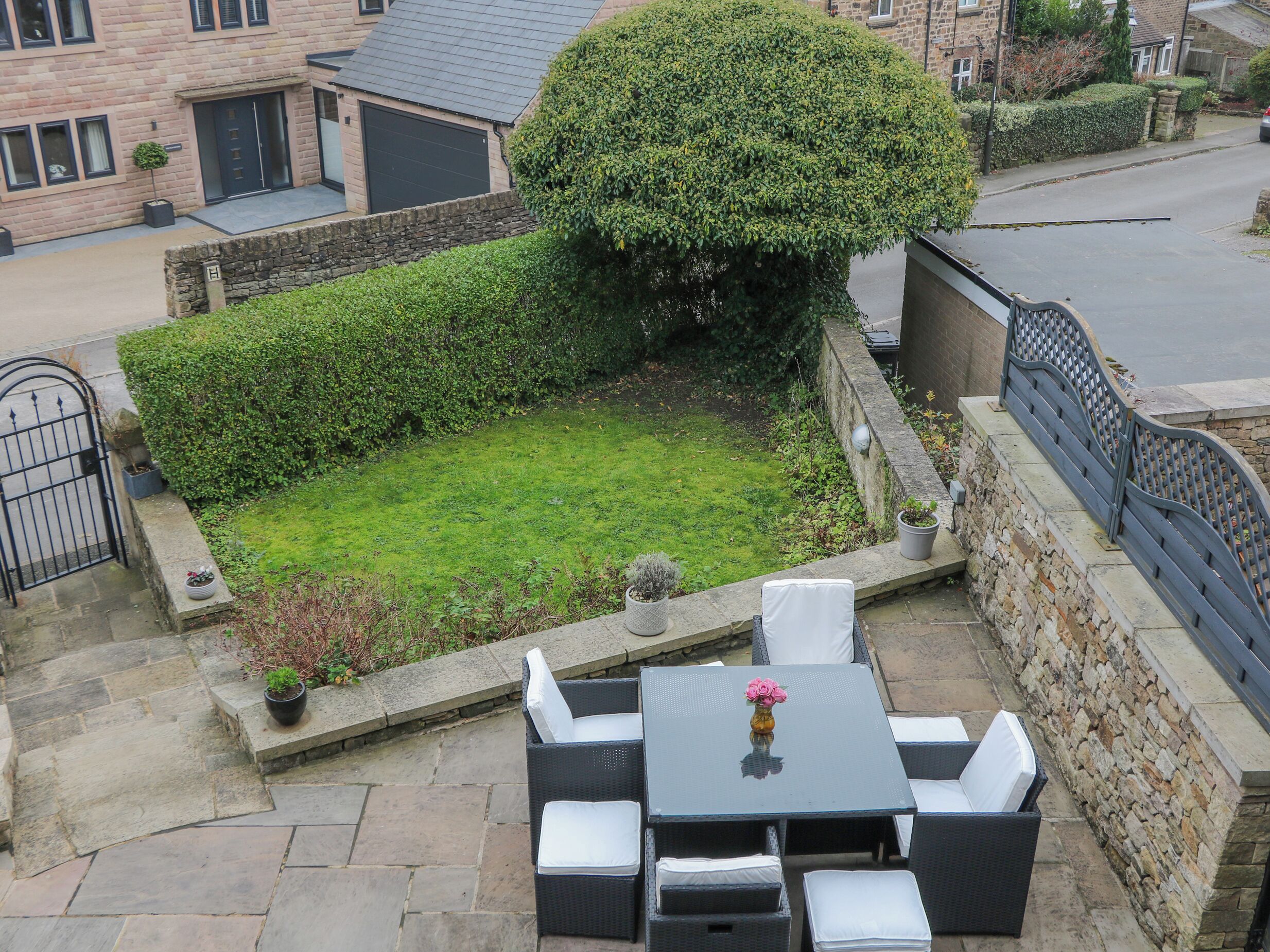 Eine Terrasse mit Tisch und Stühlen, ein kleiner Garten mit Hecke und Baum sowie eine Steinsäule.