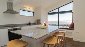 A modern kitchen with a large island, bar stools, a view of the beach, and a built-in bench with cushions. - Driftwood Cottage (Ulverston)