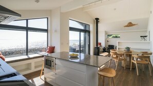A modern kitchen with a large island, a dining area with wooden chairs, and a view of the ocean. - Driftwood Cottage (Ulverston)